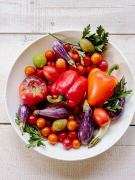 A white bowl filled with colorful vegetables including red and orange bell peppers, striped eggplants, tomatoes, cherry tomatoes, and green herbs on a light wooden surface.
