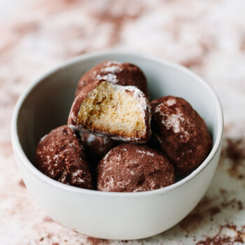A white bowl filled with chocolate snowball cookies, each dusted with cocoa powder. One cookie is broken in half to reveal its light, crumbly interior. The bowl rests on a surface sprinkled with extra cocoa powder.