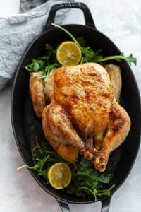 A whole roasted chicken sits in a black oval baking dish, garnished with fresh arugula and halved lemons. A gray cloth napkin rests nearby on the light-colored surface, setting for serving this delicious roasted chicken dish.