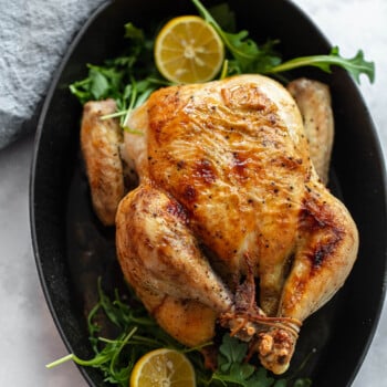 A whole roasted chicken sits in a black oval baking dish, garnished with fresh arugula and halved lemons. A gray cloth napkin rests nearby on the light-colored surface, setting for serving this delicious roasted chicken dish.