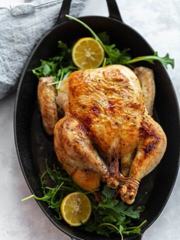 A whole roasted chicken sits in a black oval baking dish, garnished with fresh arugula and halved lemons. A gray cloth napkin rests nearby on the light-colored surface, setting for serving this delicious roasted chicken dish.