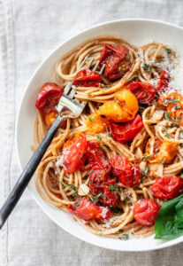 A bowl of cherry tomato pasta topped with roasted cherry tomatoes, grated cheese, herbs, and a fresh basil leaf, with a fork resting in the pasta.