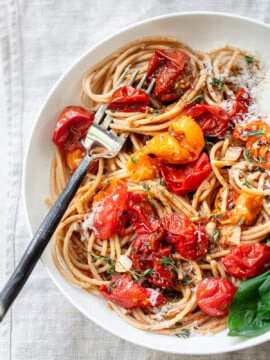A bowl of cherry tomato pasta topped with roasted cherry tomatoes, grated cheese, herbs, and a fresh basil leaf, with a fork resting in the pasta.