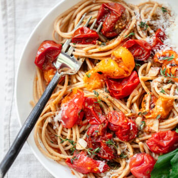 A bowl of cherry tomato pasta topped with roasted cherry tomatoes, grated cheese, herbs, and a fresh basil leaf, with a fork resting in the pasta.