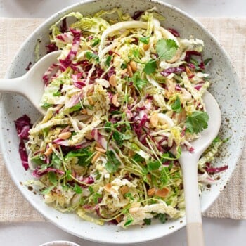 A ceramic salad bowl on a napkin, filled with green cabbage and radicchio slaw, with serving soons.