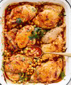 Harissa chicken thighs with a golden, seasoned crust, garnished with fresh herbs, surrounded by chickpeas and a rich, spiced red sauce, served in a white roasting pan with a spoon scooping some chickpeas.