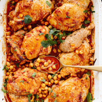 Harissa chicken thighs with a golden, seasoned crust, garnished with fresh herbs, surrounded by chickpeas and a rich, spiced red sauce, served in a white roasting pan with a spoon scooping some chickpeas.