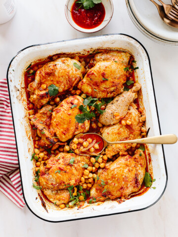 A white roasting pan filled with harissa chicken thighs, chickpeas, and tomato sauce, garnished with fresh herbs. A serving spoon rests in the pan. A small bowl of sauce and stacked plates are nearby on a white surface.