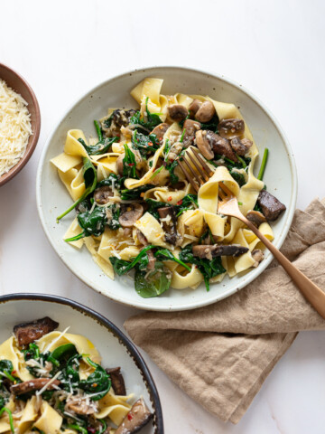 White bowls serving pappardelle pasta with spinach mushroom sauce, with a linen napkin and grated cheese on the side.