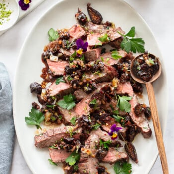 An image of an oval serving platter containing sliced grilled steak topped with a fig and parsley sauce, with lime slices.