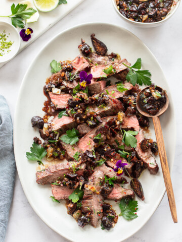 An image of an oval serving platter containing sliced grilled steak topped with a fig and parsley sauce, with lime slices.