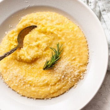 A white bowl filled with creamy polenta, topped with grated cheese and a sprig of rosemary. An ornate spoon rests in the creamy yellow mixture, while a light cloth napkin is partially visible in the background.