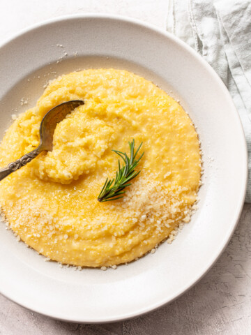 A white bowl filled with creamy polenta, topped with grated cheese and a sprig of rosemary. An ornate spoon rests in the creamy yellow mixture, while a light cloth napkin is partially visible in the background.