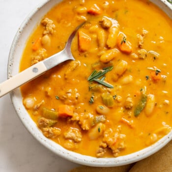 A bowl of thick orange turkey pumpkin chili with chunks of meat, carrots, celery, white beans, and herbs, served with a spoon and garnished with fresh rosemary on a white surface with a brown napkin nearby.