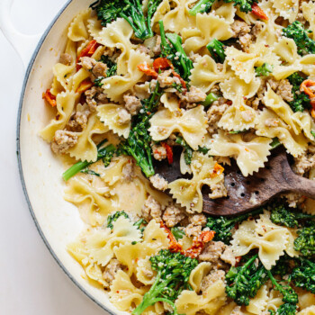 A pan filled with bow tie pasta, broccoli, ground sausage, and sun-dried tomatoes, all mixed in a creamy sauce, with a wooden spoon resting in the dish.