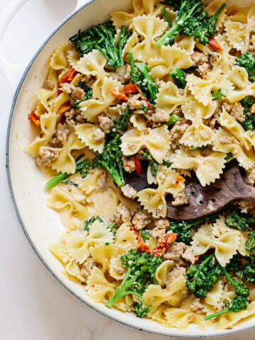 A pan filled with bow tie pasta, broccoli, ground sausage, and sun-dried tomatoes, all mixed in a creamy sauce, with a wooden spoon resting in the dish.