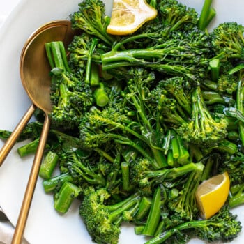 A white bowl filled with sautéed broccolini recipe, topped with two lemon wedges, with two gold serving utensils resting inside the bowl.
