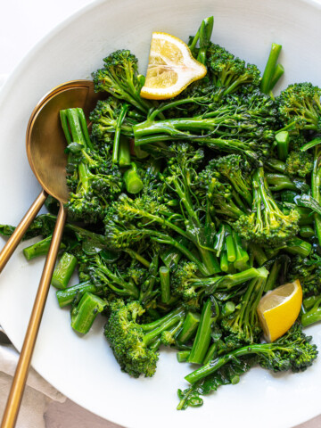 A white bowl filled with sautéed broccolini recipe, topped with two lemon wedges, with two gold serving utensils resting inside the bowl.