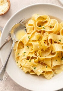 A bowl of wide pappardelle lemon pasta is topped with a creamy sauce, grated cheese, fresh lemon zest, and chopped parsley. A fork and spoon rest on the side of the bowl.