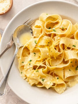 A bowl of wide pappardelle lemon pasta is topped with a creamy sauce, grated cheese, fresh lemon zest, and chopped parsley. A fork and spoon rest on the side of the bowl.