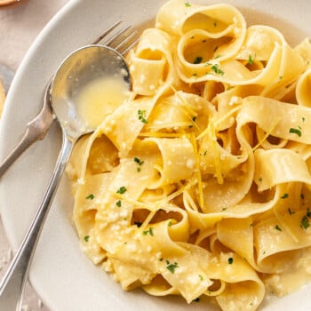 A bowl of wide pappardelle lemon pasta is topped with a creamy sauce, grated cheese, fresh lemon zest, and chopped parsley. A fork and spoon rest on the side of the bowl.