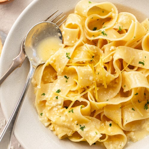 A bowl of wide pappardelle lemon pasta is topped with a creamy sauce, grated cheese, fresh lemon zest, and chopped parsley. A fork and spoon rest on the side of the bowl.