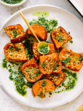 Roasted honeynut squash chunks topped with herbed breadcrumbs and green sauce are arranged on a white plate, with extra green sauce drizzled and a small yellow spoon resting in the dish.