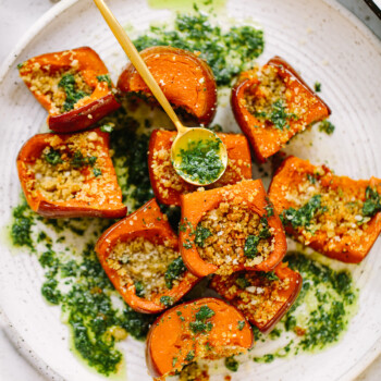 Roasted honeynut squash chunks topped with herbed breadcrumbs and green sauce are arranged on a white plate, with extra green sauce drizzled and a small yellow spoon resting in the dish.