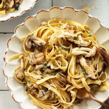 A bowl of mushroom pasta fettuccine mixed with sautéed mushrooms and garnished with grated cheese and herbs, a fork twirling the pasta nearby, and a bowl of extra cheese in the background.