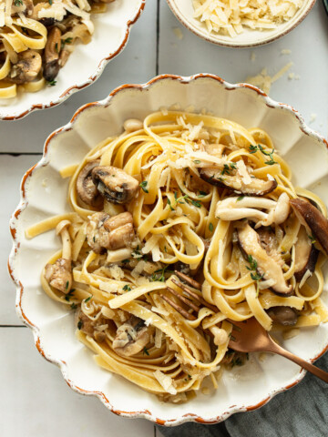 A bowl of mushroom pasta fettuccine mixed with sautéed mushrooms and garnished with grated cheese and herbs, a fork twirling the pasta nearby, and a bowl of extra cheese in the background.