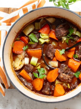 A pot of French beef stew with chunks of beef, carrots, celery, leeks, and garnished with fresh parsley. The rich and colorful stew features a ladle resting inside the pot and a striped cloth beside it.