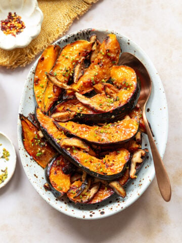 Glazed acorn squash slices roasted and paired with shiitake mushrooms and garnished with fresh herbs and spices, served on an oval plate with a spoon. Small bowls with seasonings are nearby on a light surface.