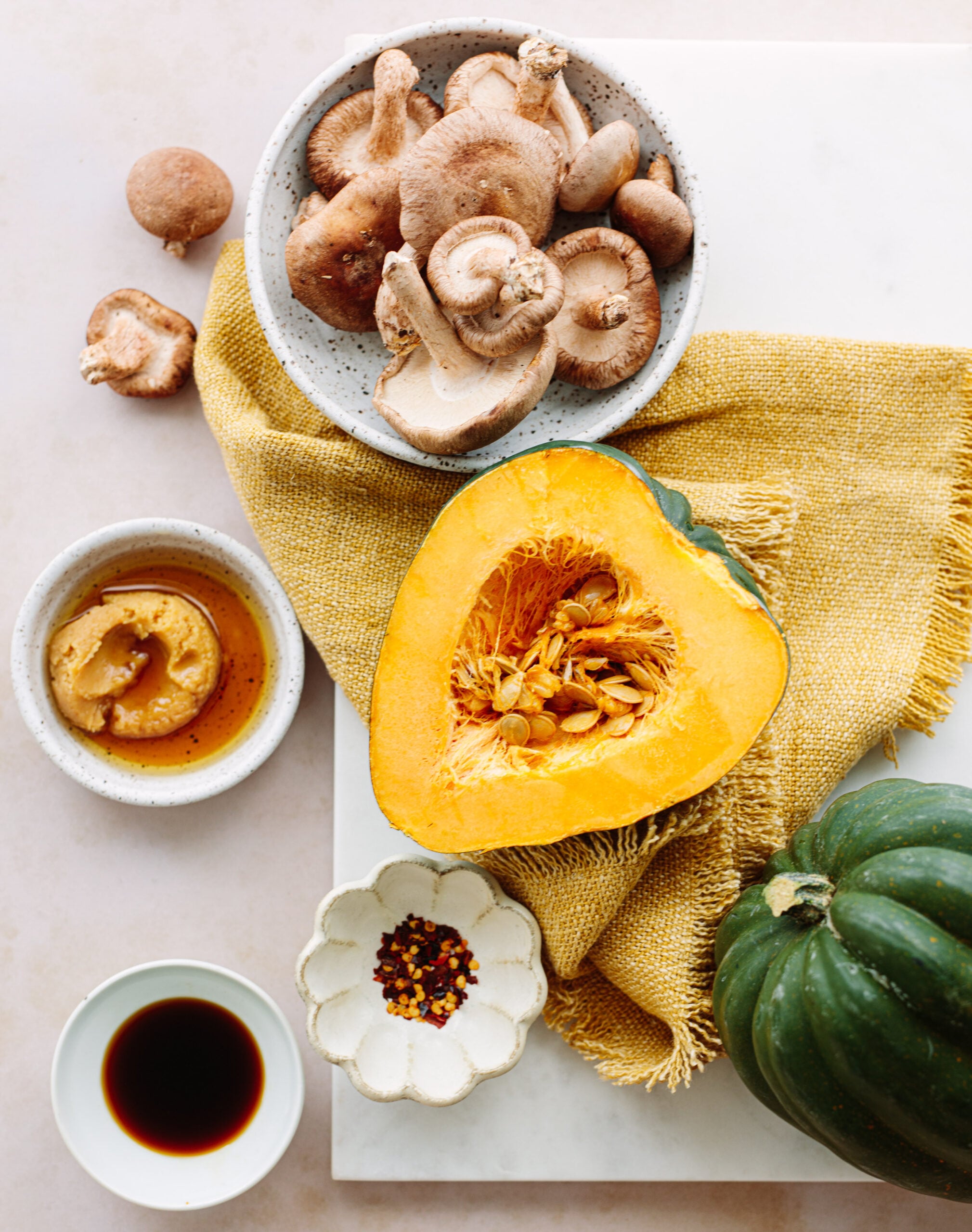 A halved glazed acorn squash, shiitake mushrooms in a bowl, soy sauce, red pepper flakes, and miso paste are arranged on a white surface with a yellow cloth.