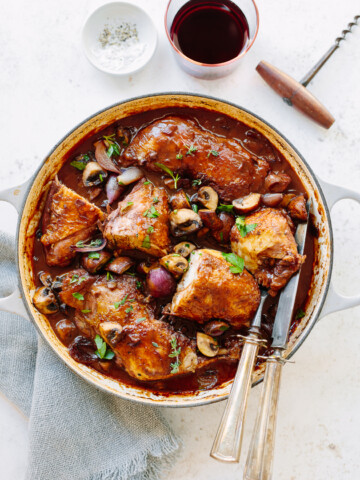 A pot of coq au vin with browned chicken, mushrooms, and onions in rich red wine sauce, garnished with parsley. Nearby are a glass of red wine, a corkscrew, a small bowl of salt, and a gray napkin.