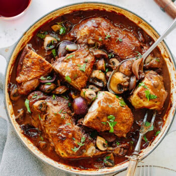 A pot of coq au vin with tender chicken, mushrooms, onions, and herbs in a rich brown sauce is served with a spoon and fork; two glasses of red wine and a bowl of salt are nearby.