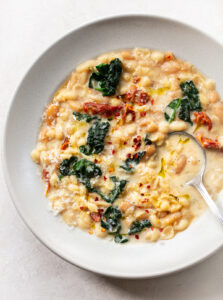 A bowl of creamy pasta fagioli-inspired white bean soup with kale, sun-dried tomatoes, grated cheese, red pepper flakes, and olive oil, served with a spoon on a light background.