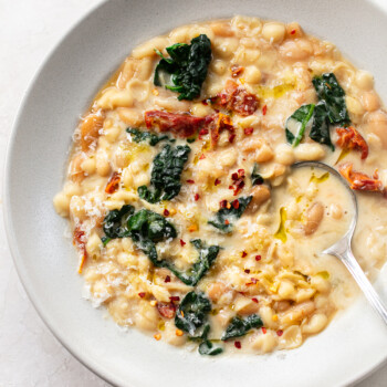 A bowl of creamy pasta fagioli-inspired white bean soup with kale, sun-dried tomatoes, grated cheese, red pepper flakes, and olive oil, served with a spoon on a light background.
