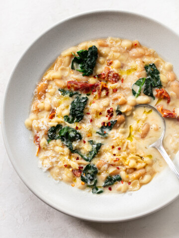 A bowl of creamy pasta fagioli-inspired white bean soup with kale, sun-dried tomatoes, grated cheese, red pepper flakes, and olive oil, served with a spoon on a light background.