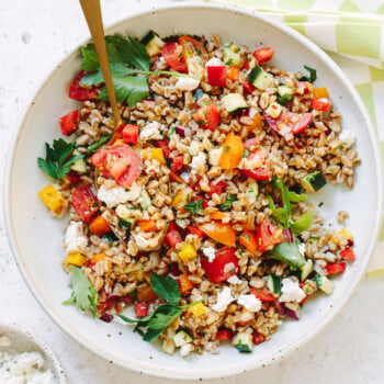A bowl of farro salad with chopped tomatoes, cucumbers, yellow bell peppers, fresh parsley, and crumbled cheese, served with a gold spoon on a white surface next to a green and white checkered napkin.