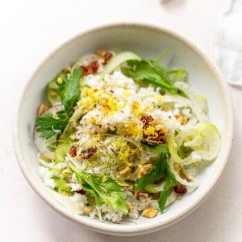 A celery salad with walnuts, shredded cheese, garnished with fresh parsley and fennel, on a light background.