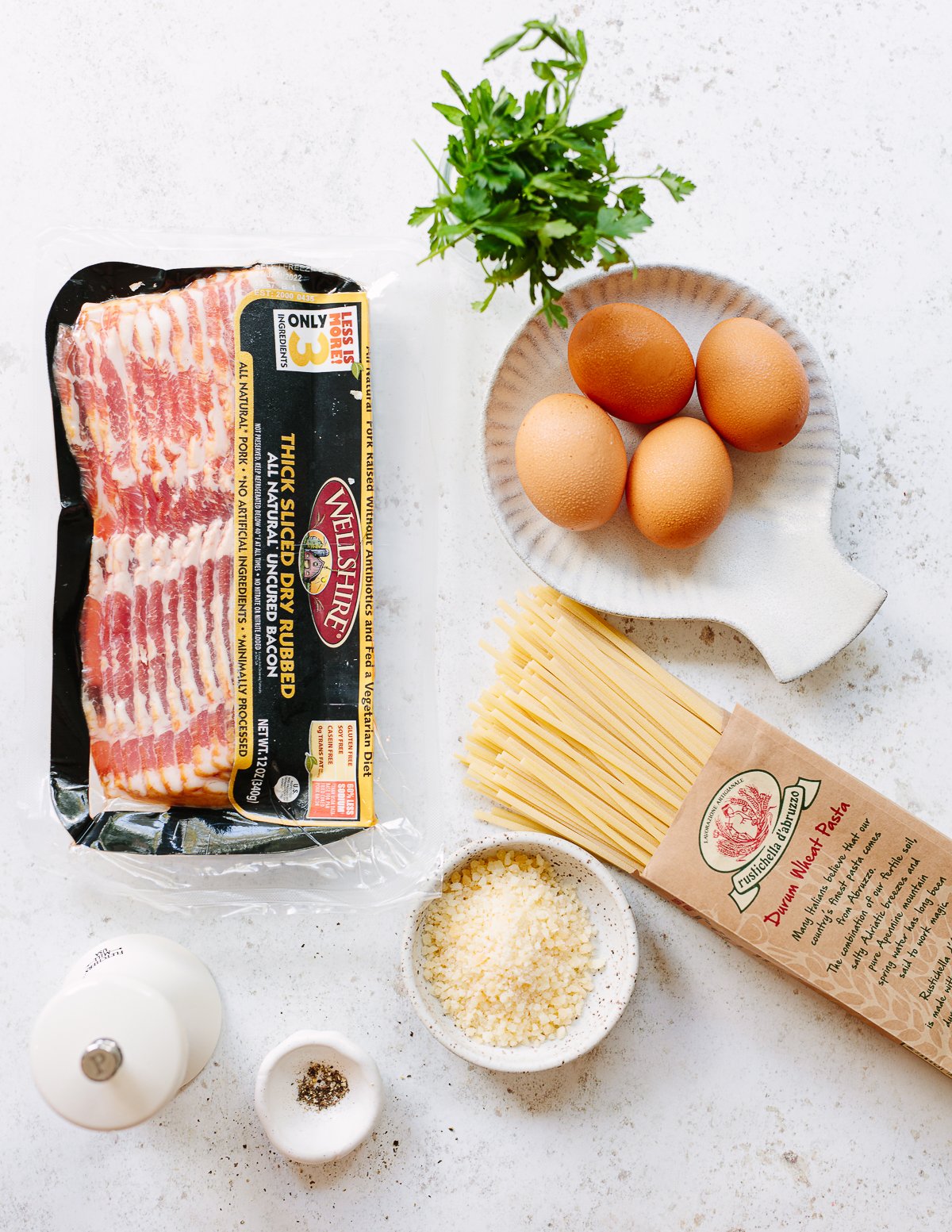 Photo showing ingredients to make pasta carbonara, with a package of bacon, eggs, grated cheese, black pepper, pasta noodles and parsley.