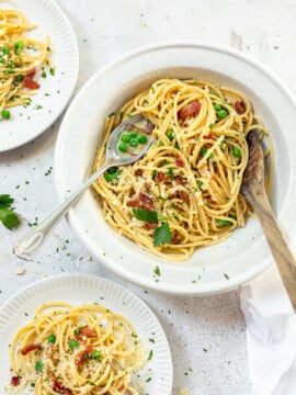 A white bowl of spaghetti carbonara with green peas, bacon bits, and parsley. Shavings of cheese and a serving spoon are on top. Two smaller plates with similar servings are nearby on a light textured surface.