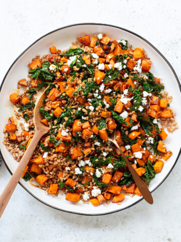 A white plate filled with sweet potato farro salad—roasted sweet potatoes, leafy greens, grains, and crumbled feta cheese—with two wooden serving utensils resting inside.
