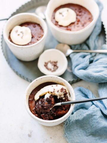 Three ceramic cups filled with rich chocolate pudding cake, each topped with cream and chocolate shavings, sit on a round tray with a blue cloth and a small dish of salt beside them. A spoon rests in the foreground cup.