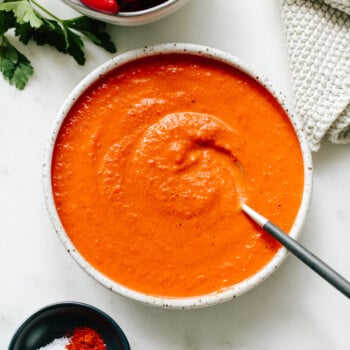A bowl of creamy romesco sauce with a spoon, surrounded by a bowl of roasted red peppers, a small dish of salt and paprika, and a textured cloth on a white surface.
