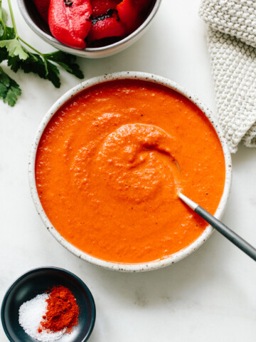 A bowl of creamy romesco sauce with a spoon, surrounded by a bowl of roasted red peppers, a small dish of salt and paprika, and a textured cloth on a white surface.