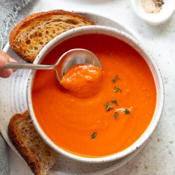 A bowl of creamy tomato soup garnished with fresh herbs, served with two slices of toasted bread; a hand lifts a spoonful of the rich, creamy tomato soup above the bowl. A small dish of salt and pepper sits nearby.
