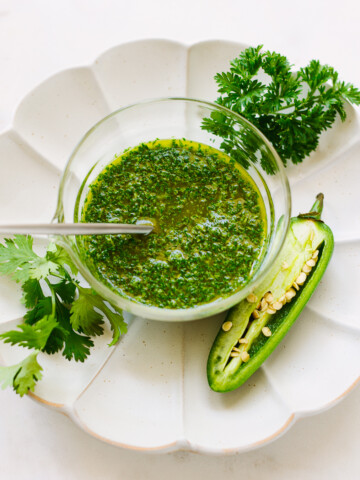 A condiment bowl full of green herb sauce, with a spoon, sliced jalapeno pepper and herb sprigs on the side.