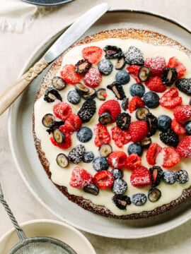A round almond cake topped with creamy white icing, fresh raspberries, blueberries, blackberries, and sliced grapes, dusted with powdered sugar, sits on a plate with a knife beside it.