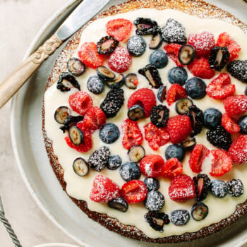 A round almond cake topped with creamy white icing, fresh raspberries, blueberries, blackberries, and sliced grapes, dusted with powdered sugar, sits on a plate with a knife beside it.
